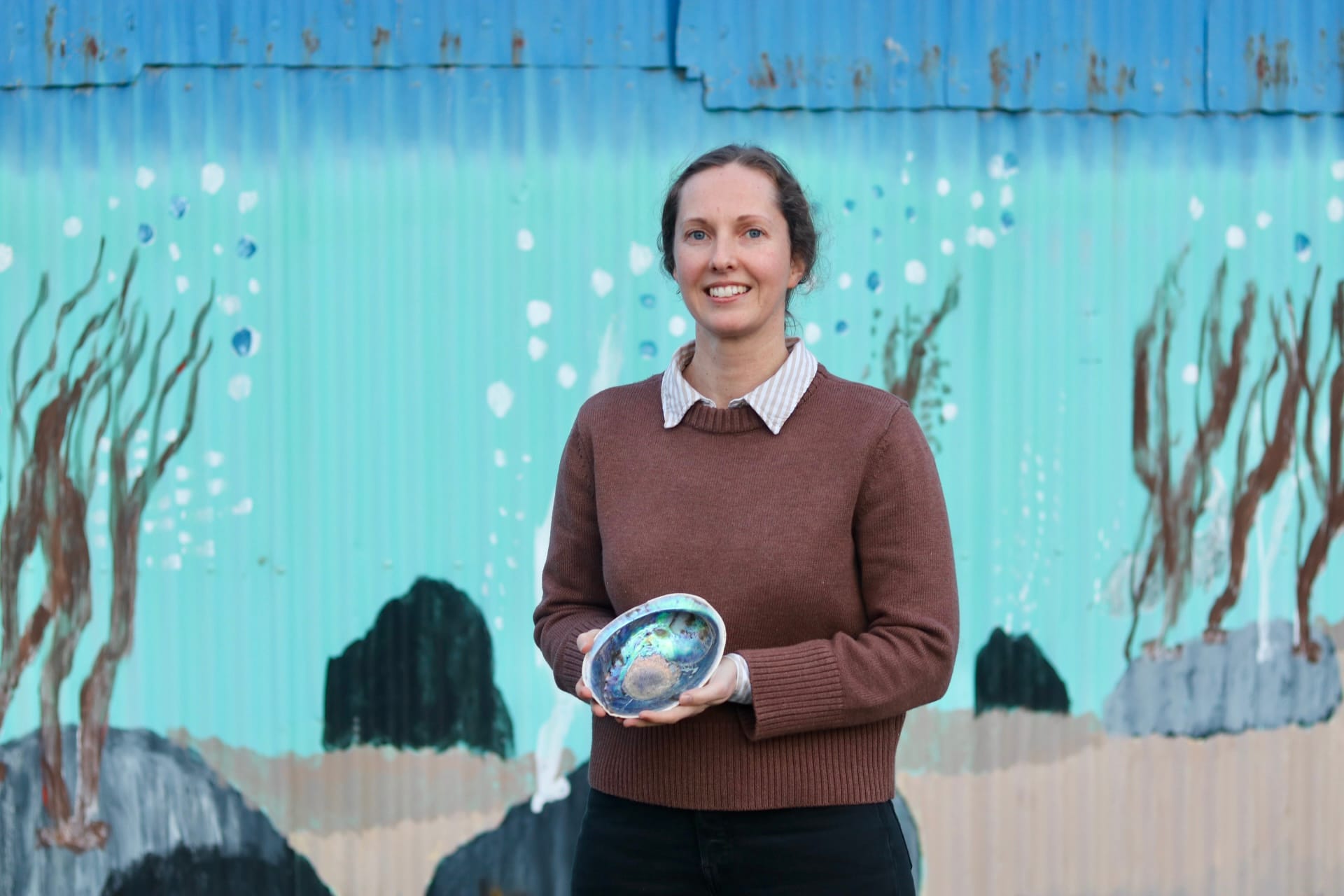 Ocean Shell owner Nina Shields holding a polished paua shell in front of a painted underwater mural.